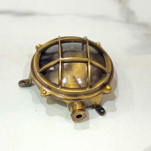 May include: A round, antique-style brass bulkhead light fixture featuring a protective metal cage over a clear glass dome. The fixture has a circular frame with visible screw fasteners and a small, protruding cylindrical conduit entry point at the base.