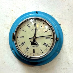 May include: A round, blue wall clock with a cream-colored face and black Roman numerals. The clock face features the text "SMITHS LONDON" and a ship illustration. The clock has a metal frame and a hinged door.