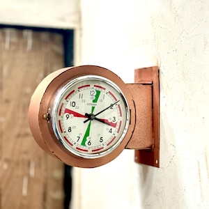 May include: A vintage, double-sided clock with a copper-colored metal casing. The clock face is white with black numerals and red and green accents. The clock is mounted on a wall.