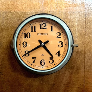 May include: A round Seiko clock with a light orange face and black numerals. The clock has a silver-colored bezel and black hands. The clock is mounted on a wooden surface. The text "SEIKO" is printed on the clock face.