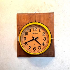 May include: A vintage Seiko wall clock with a yellow frame and a light orange face. The clock is mounted on a rectangular wooden board. Black hands and numerals are visible on the clock face. The clock is hanging on a white wall.