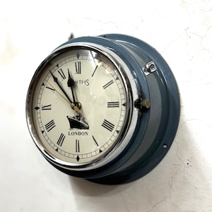 Könnte beinhalten: Eine runde, blaue Wanduhr mit silbernem Rand. Das cremefarbene Zifferblatt hat schwarze römische Ziffern und Zeiger. Das Zifferblatt trägt die Aufschrift "LONDON" und eine Schiffsillustration. Die Uhr ist an einer weißen Wand befestigt.