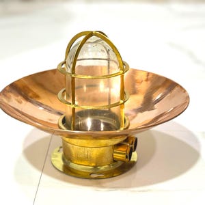 Puede incluir: Una lámpara náutica de latón y cobre de estilo vintage. La lámpara presenta un globo de cristal transparente encerrado en una jaula de latón, colocado sobre un reflector en forma de cuenco de color cobre. La base es de latón con tres pequeños puertos.