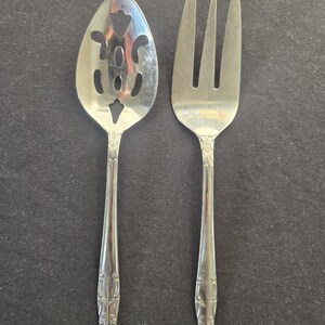 May include: A silver-toned metal serving spoon and a matching serving fork. The spoon features a bowl with decorative cutouts, while the fork has three long tines. Both utensils have ornate floral engravings on the handles and a flared base.