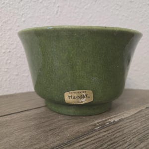 May include: A small, round, moss-green ceramic bowl with a flared rim and a pedestal base. A tan-colored sticker is affixed to the front of the bowl, featuring the text "COPYRIGHTED Haeger" in dark brown lettering.
