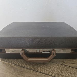 May include: A vintage Samsonite briefcase in shades of brown and black. The briefcase has a textured exterior, a brown handle, and a silver-toned metal accent. The Samsonite logo is visible on the front.