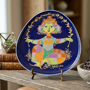 May include: A rounded triangular ceramic plate with a deep blue background featuring a colorful, stylized illustration of a seated figure in shades of green, orange, and purple. The design includes gold-colored line art accents and a signature.