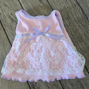 Op de afbeelding: Een mouwloze lichtroze top met een witte kanten overlay met bloemmotief op het onderste gedeelte. Het kledingstuk is afgewerkt met een lichtpaarse lintrand langs de halslijn en armsgaten, en een bijpassende lichtpaarse strik in het midden.
