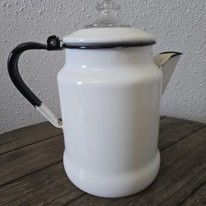 May include: White enamel coffee pot with a black handle and trim. The pot has a clear glass lid and a spout for pouring. The vintage kitchenware is likely used for brewing coffee or other hot beverages.