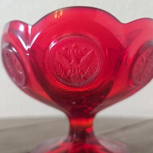 Puede incluir: Un cuenco de vidrio rojo brillante con borde festoneado y base de pedestal. El cuenco presenta diseños circulares en relieve con un emblema de águila. El vidrio tiene un acabado brillante, reflejando la luz. Objeto decorativo.