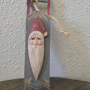 May include: A hand-carved wooden ornament shaped like a sled, featuring a painted Santa Claus face with a red hat and long white beard. The sled is painted grey with red runners and includes a natural jute twine loop for hanging purposes.