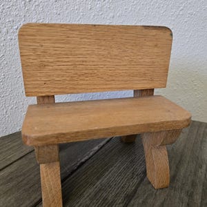 May include: A small, handcrafted wooden bench with a backrest. The bench is made of light-colored wood and has a simple, rustic design. The bench is suitable for decorative purposes or for small dolls.