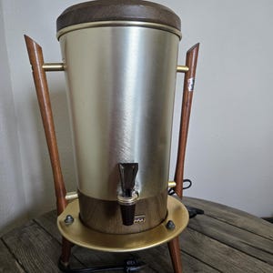 May include: Vintage coffee percolator with a brushed metal body, wooden lid, and legs. The percolator has a brass-colored base and accents, and a black spout. A classic kitchen appliance.