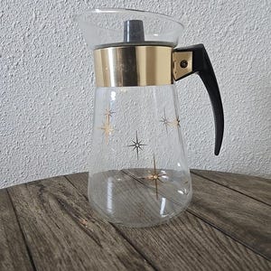 May include: A vintage glass coffee carafe with a gold-colored band and black handle. The carafe features a clear glass body with gold starburst designs. The carafe is likely from the mid-century era.