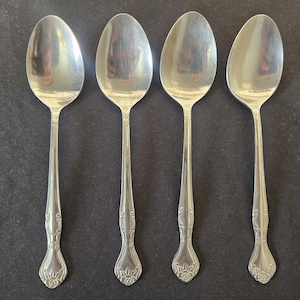 May include: A set of four silver-toned metal spoons arranged in a row. Each spoon features a smooth, oval-shaped bowl and a slender handle that widens toward the base, decorated with an embossed floral motif at the very tip of the handle.
