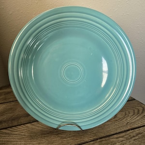Puede incluir: Un plato redondo de cerámica de color turquesa claro que presenta una serie de crestas circulares concéntricas en relieve a lo largo del borde y un patrón de anillos a juego centrado en el medio, creando una superficie brillante.