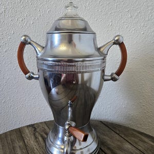 May include: A vintage silver-toned coffee percolator with brown handles and a spigot. The percolator has a glass lid and sits on a wooden surface. The design is classic and elegant, suitable for serving coffee.