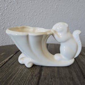 May include: A white ceramic planter in the shape of a cornucopia, with a squirrel figure on the side. The planter has a curved design and sits on a wooden surface.
