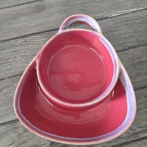 May include: A vibrant pink ceramic serving dish with a handle. The dish has a rounded shape with a slightly iridescent rim. The handle is arched, and the dish sits on a matching, wide base.