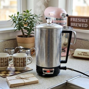 May include: A silver metal electric percolator with a black handle and base, featuring a clear glass knob on the lid and a power cord. The base has a black label with the text FREFEENGNR and a small orange indicator light that is illuminated.