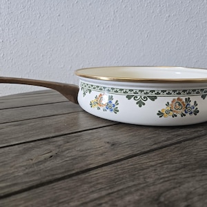 May include: White enamel pan with a wooden handle. The pan features a gold rim and a decorative band with green and floral patterns. The floral design includes blue, yellow, and orange flowers. The pan is set on a wooden surface.