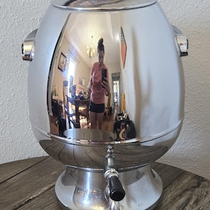 May include: A large, shiny, chrome-plated coffee urn with a glass lid and a spigot. The urn has a rounded body and sits on a pedestal base. The surface reflects the surrounding environment.
