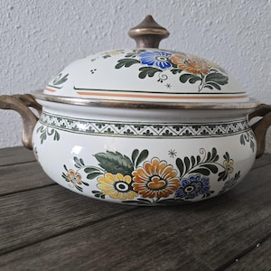 May include: A white ceramic casserole dish with a lid, adorned with colorful floral patterns in orange, yellow, and blue. The dish has gold-toned handles and a gold finial on the lid. A decorative piece for serving.