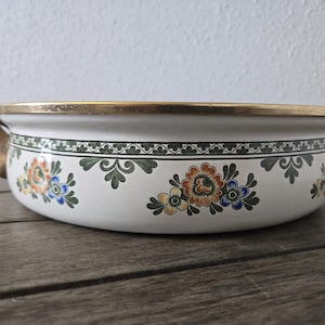 May include: A white ceramic casserole dish with a gold rim and handles. The dish features a floral design in orange, blue, and yellow, with a green and black border. The dish is on a wooden surface.