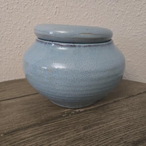 May include: A light blue ceramic jar with a matching lid, featuring a rounded, bulbous body and a subtle crackle glaze finish. The vessel has a smooth, curved silhouette and a slightly darker rim where the lid meets the base of the container.