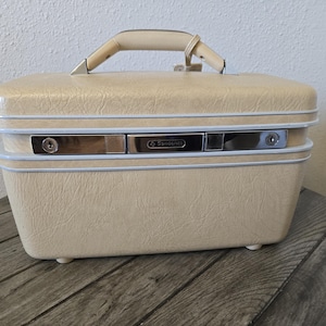 Pode incluir: Uma mala de viagem Samsonite vintage creme com acabamento texturizado. Possui uma alça creme, fechos prateados e uma moldura azul claro. A mala retangular assenta em pequenos pés.