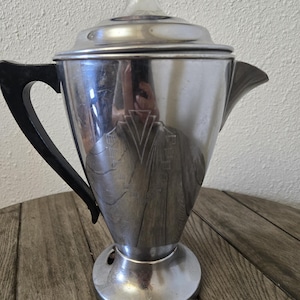 May include: A polished silver-tone metal percolator coffee pot featuring a black handle, a clear glass knob on the lid, and a tapered body with an etched geometric design on the front. The coffee pot has a wide base and a prominent pouring spout.
