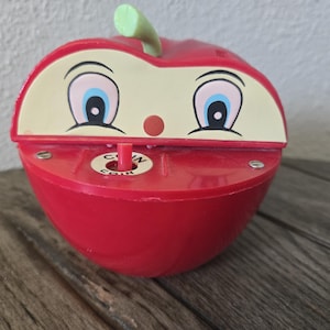 May include: A red plastic apple-shaped coin bank with a cartoon face featuring large blue and white eyes and a small red nose. A green stem sits on top, and a small white circular slot labeled COIN is positioned in the center of the red base.