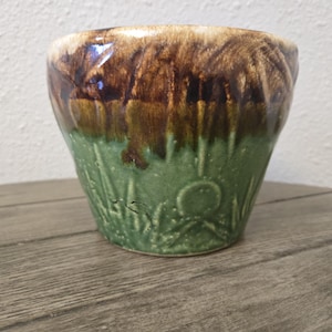 May include: A ceramic planter featuring a two-tone glaze with a dark brown drip pattern at the rim transitioning into a textured sage green base. The lower portion of the pot displays a raised relief design of blades of grass and a small circular element.