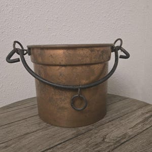 Puede incluir: Un cubo cilíndrico de cobre con una pátina oscura y desgastada. Cuenta con un asa giratoria de hierro oscuro sujeta a dos bucles laterales, con un pequeño anillo de hierro adicional que cuelga del centro de la parte inferior curva del asa.