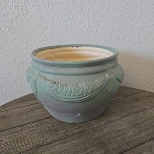 Peut inclure: Un pot de fleurs en céramique vert sauge avec une finition mate et texturée. L'extérieur présente un motif en relief de guirlandes de roses et de festons entourant le milieu. L'intérieur du récipient en forme de bol est blanc crème lisse.