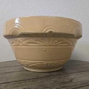 May include: A light tan ceramic mixing bowl with a wide rim and a thick, raised band featuring a repeating semi-circular pattern. The exterior body of the bowl is decorated with horizontal bands and embossed sunburst or fan-like geometric motifs.