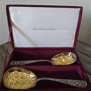 Könnte beinhalten: Ein Set aus zwei Servierlöffeln in einer bordeauxroten Samtbox. Die Löffel haben verzierte Silbergriffe und goldfarbene Schalen mit einem detaillierten Blumendesign. Die Box ist mit weißem Satin gefüttert und trägt den Text "American Silversmiths Collection".
