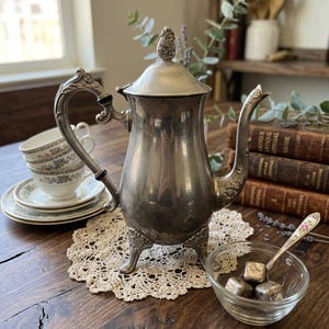 Puede incluir: Una tetera de metal plateado con tapa, asa curva y pico, apoyada sobre tres patas ornamentadas. La tetera presenta detalles florales decorativos en el asa, el pico y el remate de la tapa. Está colocada sobre un tapete de encaje redondo de color crema.