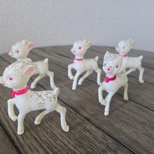 May include: A set of four white plastic deer figurines, each featuring a bright pink collar and subtle silver glitter accents on the back and head. The deer are depicted in a standing pose with one front leg raised, showcasing pink painted eyes.
