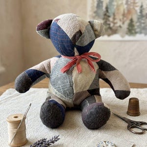 Peut inclure: Un ours en peluche en patchwork fabriqué à partir de divers morceaux de tissu dans des tons de bleu, gris, marron et beige. L'ours présente un ruban rose noué autour du cou, des oreilles arrondies et un corps cousu. C'est un jouet rembourré.