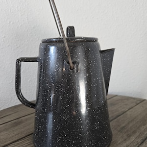 May include: A black and white speckled enamelware coffee pot with a handle and spout. The pot has a lid and a wire handle that arches over the top. The pot is sitting on a wooden surface.