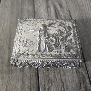Op de afbeelding: Een rechthoekige zilverkleurige metalen doos met een reliëfscène op het deksel met twee figuren in een landschap. Het deksel heeft een decoratieve rand met schelpachtige motieven en de zijkanten van de doos zijn versierd met krulwerk.