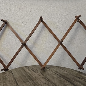 May include: A wooden expanding coat rack in a diamond pattern. The rack is made of dark brown wood and has small knobs at the ends of each arm for hanging items. It is mounted on a wall and is a decorative storage solution.