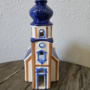 May include: A ceramic church figurine with a blue steeple and roof, white walls, and brown and blue trim. The decorative piece has arched windows and a detailed design, perfect for home decor.