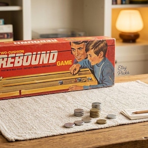 May include: A vintage Two-Cushion Rebound game box featuring red packaging with an illustration of a person playing the game. The box displays the game title and includes a set of small metal pucks and a stack of cards with a pencil on the surface.