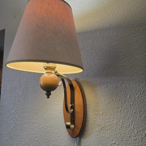 Peut inclure: Une applique murale composée d'un abat-jour conique en tissu clair, d'un élément central en bois sphérique et d'un bras métallique incurvé fixé sur une plaque arrière ovale en bois. La lampe diffuse une lueur chaude sous l'abat-jour.