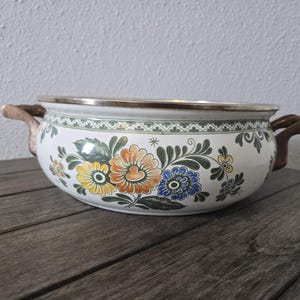 May include: A white ceramic bowl with floral designs in orange, yellow, and blue. The bowl has a silver rim and brown handles. The floral pattern includes green leaves and stems. The bowl is sitting on a wooden surface.