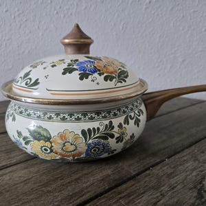 May include: A vintage, cream-colored saucepan with a lid, featuring a floral design in shades of blue, orange, and yellow. The pan has a wooden handle and a gold-colored rim. The lid is topped with a gold-colored finial.