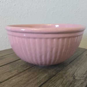 Puede incluir: Un cuenco rosa de cerámica con detalles acanalados verticales. El cuenco tiene un borde redondeado y se asienta sobre una superficie de madera. El color del cuenco es un rosa pastel suave.