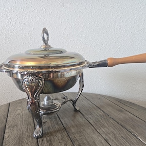 May include: A silver-plated chafing dish with a round lid featuring a central handle, a long wooden handle, and a decorative stand with three ornate legs. The base includes a small metal burner component positioned beneath the main bowl.